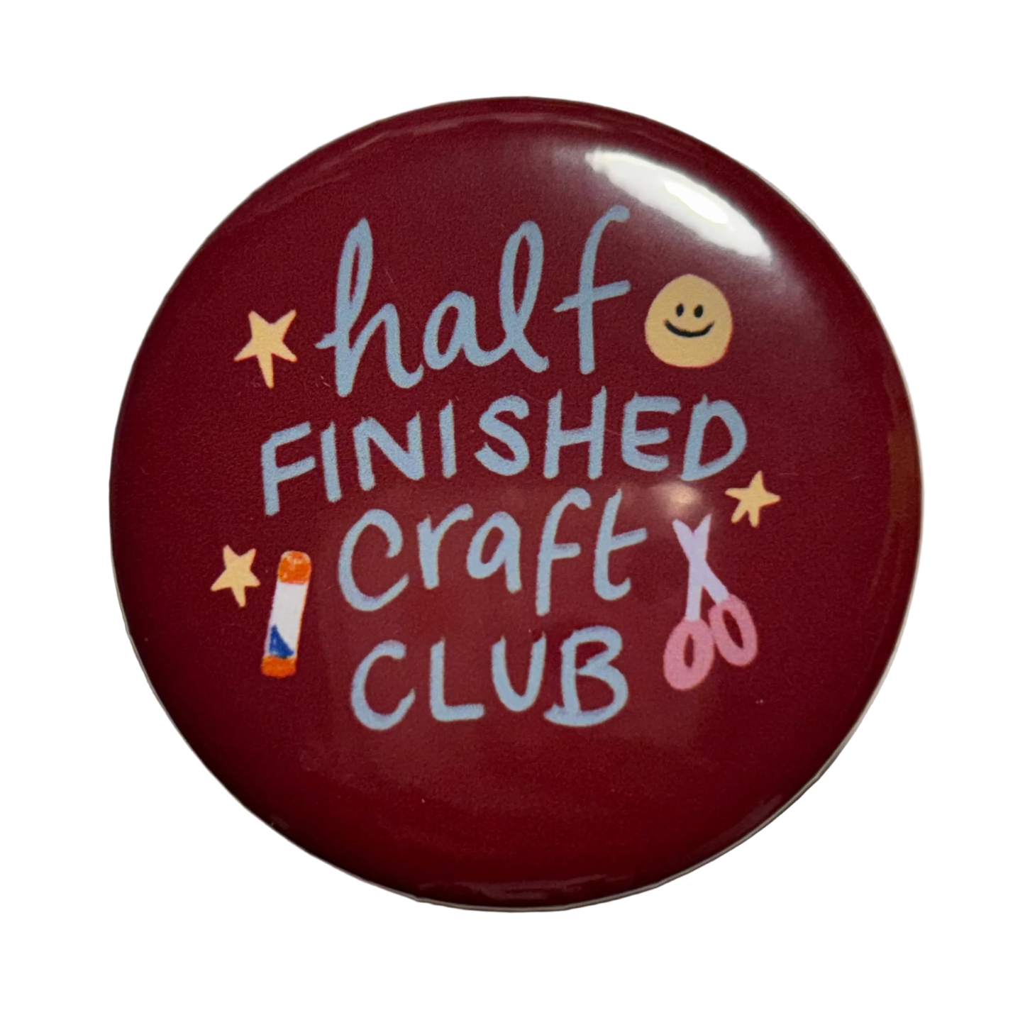 A round glossy dark red button badge featuring the text 'half FINISHED Craft CLUB' in light blue lettering, decorated with small yellow stars, a smiley face, a paintbrush, and scissors.