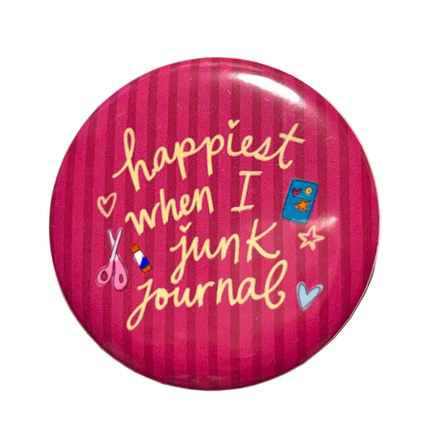 A round pinback button badge featuring a pink and magenta striped background with the phrase 'happiest when I junk journal' in yellow script, decorated with small craft-themed icons including scissors, a blue notebook, and hearts.