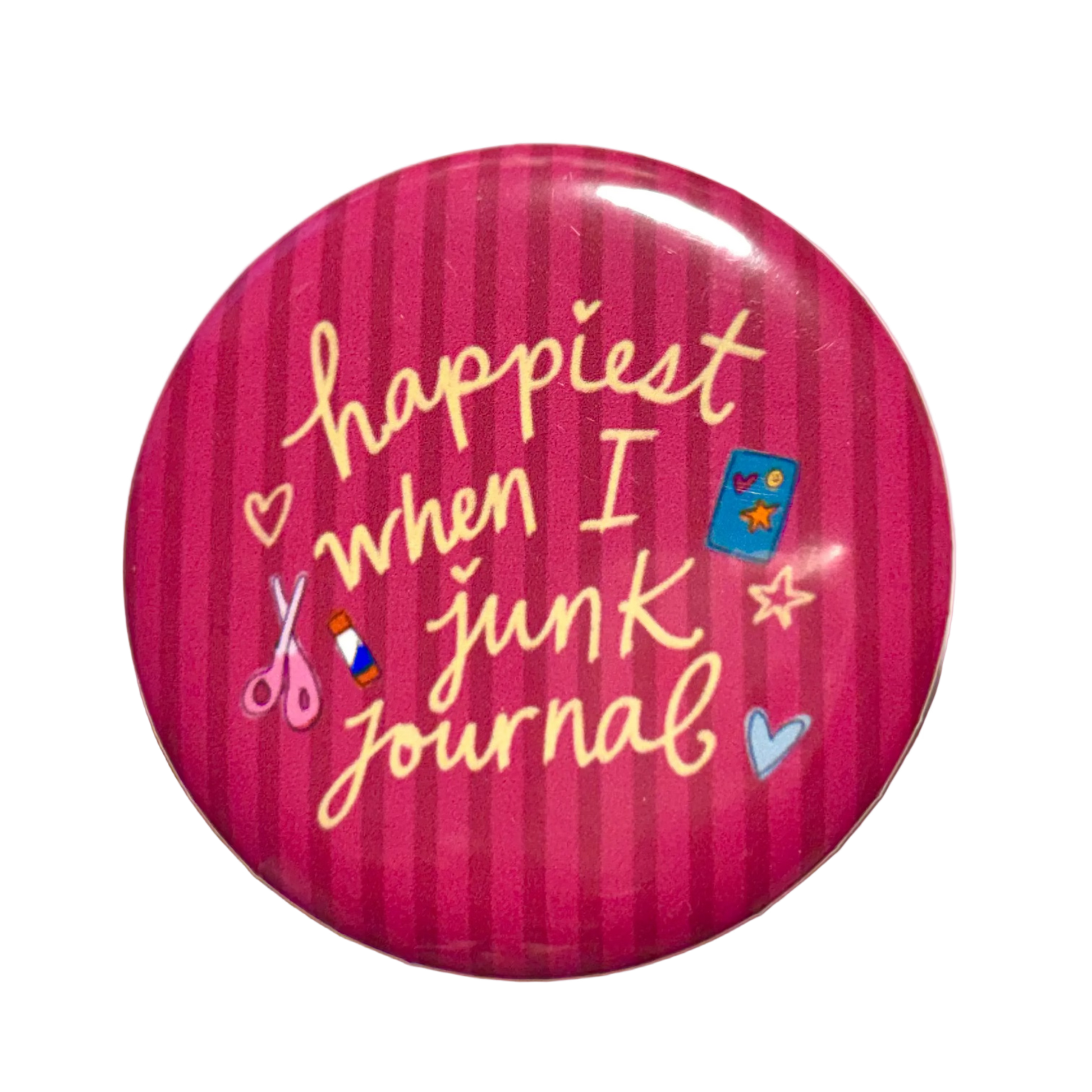 A round pinback button badge featuring a pink and magenta striped background with the phrase 'happiest when I junk journal' in yellow script, decorated with small craft-themed icons including scissors, a blue notebook, and hearts.