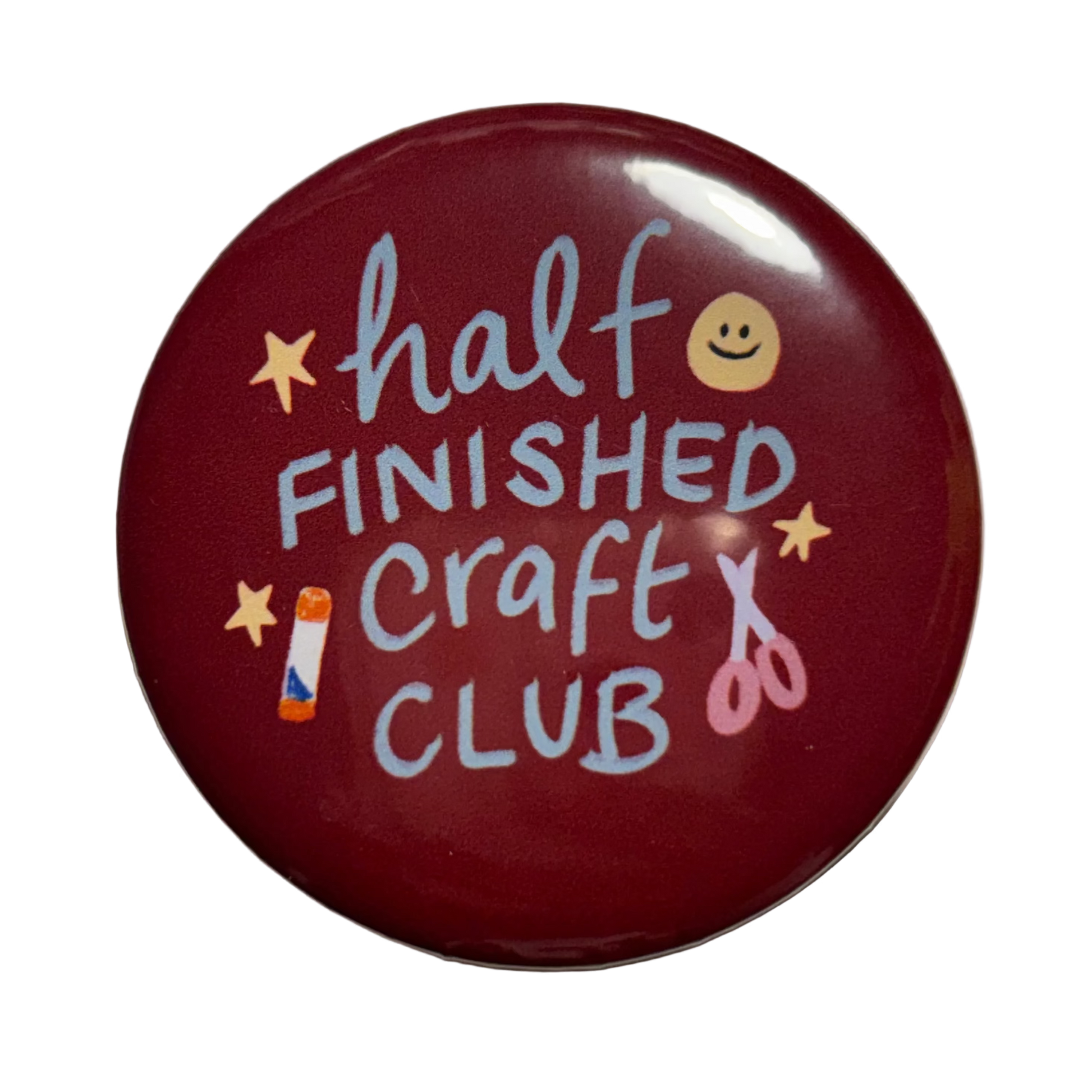 A round glossy dark red button badge featuring the text 'half FINISHED Craft CLUB' in light blue lettering, decorated with small yellow stars, a smiley face, a paintbrush, and scissors.