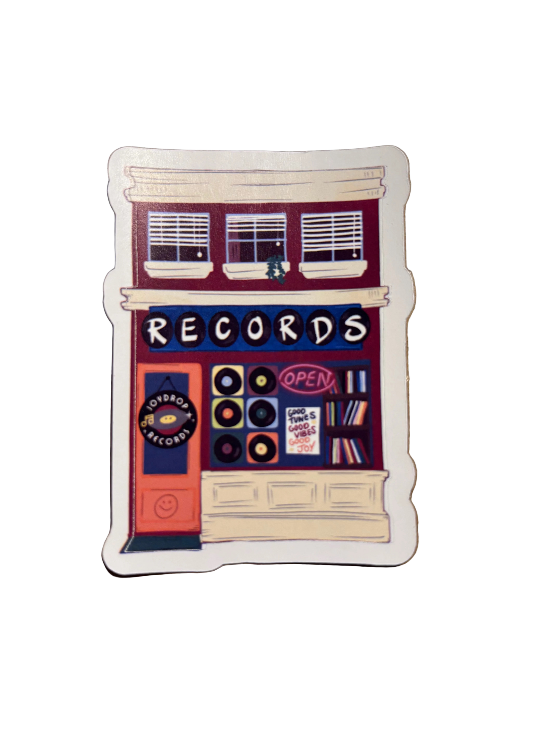 A die-cut sticker featuring an illustrated record store with vinyl records, windows, and signs including 'RECORDS' and 'OPEN', placed on a brown textured surface.